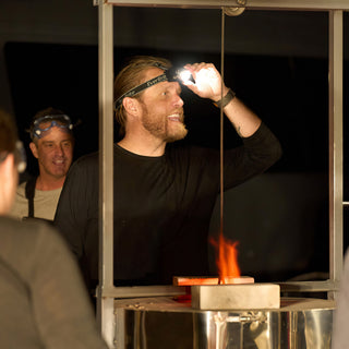 A potter adjusts a bright headlamp beside a metal frame with a pulley cord and a small flame below at The Clay Hole in Draper, Utah