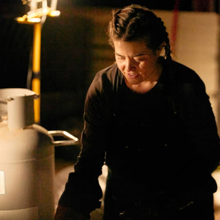 A potter with braided hair and dark clothes looks down and smiles beside a propane tank at The Clay Hole in Draper, Utah