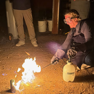 A smiling potter in goggles and gloves crouches beside a white pump sprayer as flames rise around a small vase at The Clay Hole in Draper, Utah