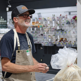 A smiling potter in a tan apron and ball cap holds a small tool beside a bag of clay at The Clay Hole in Draper, Utah