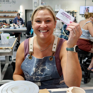A smiling potter in a denim apron holds up a small purple and white prize card at a work table at The Clay Hole in Draper, Utah