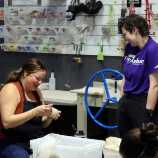 A potter laughing while turning a small unglazed bowl in their hands, with a blue-wheeled slab roller behind at The Clay Hole in Draper, Utah