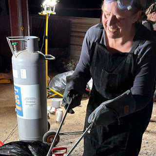 A potter in a black apron and long gloves smiles while holding tongs beside a tall gray gas cylinder at The Clay Hole in Draper, Utah