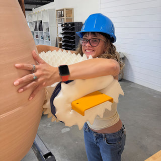 A smiling potter in a blue hard hat hugs a huge tan clay vessel while holding a cream foam piece at The Clay Hole in Draper, Utah