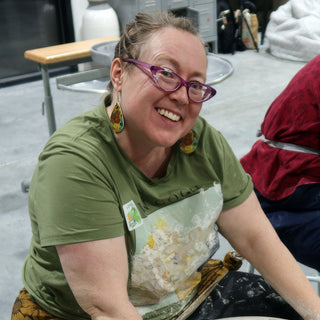 A smiling potter in a green shirt and clay-covered apron turns toward the camera wearing purple glasses at The Clay Hole in Draper, Utah