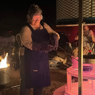 At night a potter in goggles and heavy gloves holds long tongs near a small fire in a metal bucket at The Clay Hole in Draper, Utah