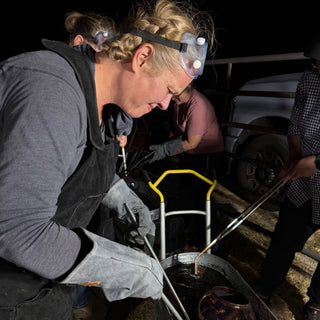 A potter in goggles and heavy gloves leans over a metal tub with tongs while another person works nearby at The Clay Hole in Draper, Utah