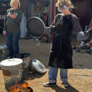 A potter in goggles, an apron and heavy gloves holds a metal lid beside trash cans with a small fire at The Clay Hole in Draper, Utah