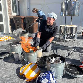 A potter in goggles and gloves kneels beside a flaming metal bucket outdoors while another stands behind with long tongs, with The Clay Hole, a pottery studio in Draper, Utah