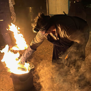 A potter in a headlamp and long gloves leans over a metal can with flames and smoke rising at night at The Clay Hole in Draper, Utah