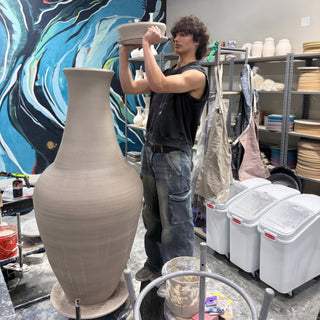 A potter holds a wide shallow dish overhead beside a tall slender vase, with a blue mural behind at The Clay Hole in Draper, Utah
