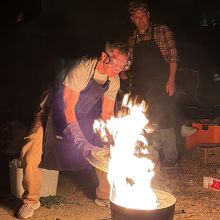 A potter in safety goggles and an apron lifts a metal lid off a barrel of tall flames at The Clay Hole in Draper, Utah