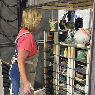 A potter in a tan apron looks over a shelf rack of glazed vases, bowls, mugs and plates at The Clay Hole in Draper, Utah