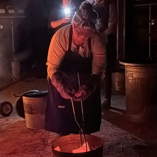 A potter in a dark apron bends over a small metal can and lowers long tongs into it at The Clay Hole in Draper, Utah