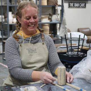 A potter with braided pigtails smiling while pressing an upright block of clay between two wooden strips at The Clay Hole in Draper, Utah