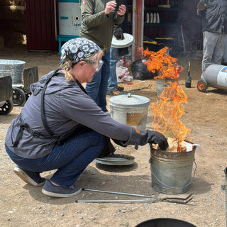 A potter in a bandana crouches and reaches a gloved hand toward flames rising from a metal bucket at The Clay Hole in Draper, Utah