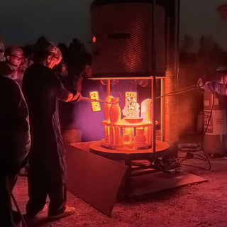 A potter reaches tongs toward glowing orange vessels on a kiln platform while a crowd watches in the dark at The Clay Hole in Draper, Utah
