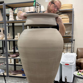 A potter lowers a wide flared lid onto a huge round-bodied gray clay jar in front of studio shelves at The Clay Hole in Draper, Utah