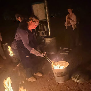 A potter in goggles and long gloves reaches tongs into a metal bucket of flames while others stand nearby at The Clay Hole in Draper, Utah