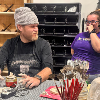 A potter in a gray knit beanie makes a scrunched face beside a container of red-handled fan brushes at The Clay Hole in Draper, Utah
