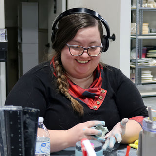 A potter wearing headphones laughs while working a small piece of clay with blue-stained fingers at a table at The Clay Hole in Draper, Utah