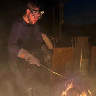 A potter in goggles, a headlamp and long gloves reaches tongs toward low flames in the dark at The Clay Hole in Draper, Utah
