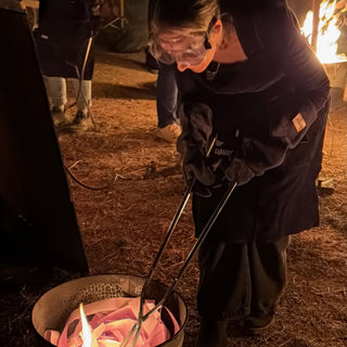 A potter in goggles and heavy gloves leans over a metal bucket of torn paper, holding long tongs at The Clay Hole in Draper, Utah