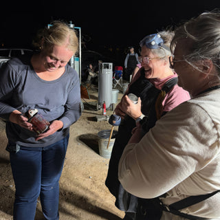 Three potters gather outdoors at night as one looks down at a small dark red lidded jar, with The Clay Hole, a pottery studio in Draper, Utah