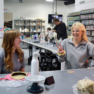 Two potters laugh at a long gray table, one flashing a peace sign, with a turntable and clay in front at The Clay Hole in Draper, Utah