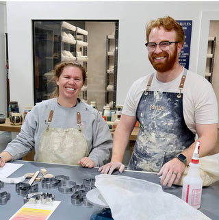 Two smiling potters pose at a worktable spread with metal cutters, wooden tools and a spray bottle at The Clay Hole in Draper, Utah