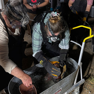 Potters in headlamps and heavy gloves lower a dark piece into a metal tub beside a cart at The Clay Hole in Draper, Utah
