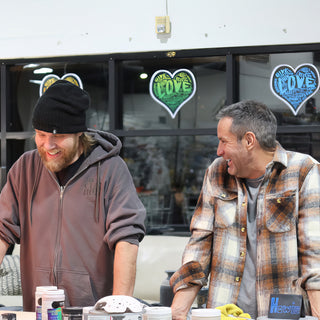 Two potters laughing together at a worktable in front of heart decals on the window, one in a black beanie at The Clay Hole in Draper, Utah