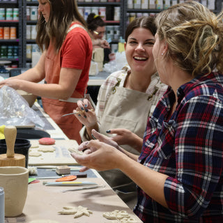 Potters laugh together at a long table covered with small clay cutouts, a wooden tool, and a pale cup at The Clay Hole in Draper, Utah