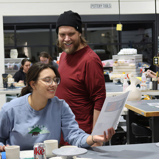 Two potters smiling at a table while looking at a printed sheet, with a pale unglazed vase nearby at The Clay Hole in Draper, Utah