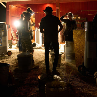 Potters in headlamps stand among metal bins and gas tanks inside a red barn lit from behind at The Clay Hole in Draper, Utah