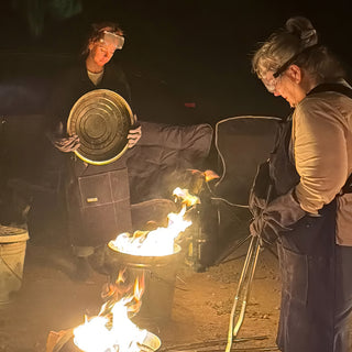 Two people in goggles and aprons tend flaming metal cans at night, one holding a lid, the other tongs at The Clay Hole in Draper, Utah