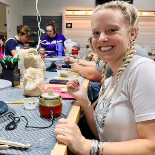 A smiling potter with braided hair sits at a long worktable with bagged clay and a small red electric device at The Clay Hole in Draper, Utah