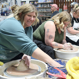 Two potters at pottery wheels, one smiling at the camera with a hand on a cone of clay at The Clay Hole in Draper, Utah
