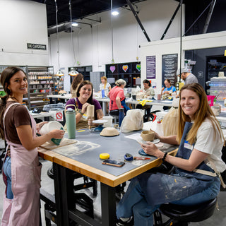 Three smiling potters work on bowls and cups at a long table with a busy class behind them at The Clay Hole in Draper, Utah