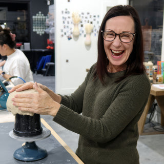 A laughing potter presses both hands onto a rounded lump of clay on a black cylinder and turntable at The Clay Hole in Draper, Utah