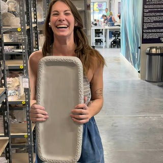 A laughing potter holds a long rectangular unglazed tray with a textured rim in front of shelves at The Clay Hole in Draper, Utah