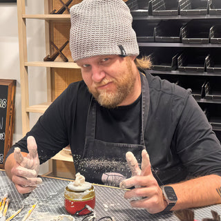 A potter in a gray beanie holding clay-covered hands up behind a small red jar topped by a figure at The Clay Hole in Draper, Utah