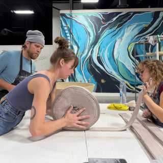 A potter rolls up a long wide strip of gray clay on a white table while others work nearby at The Clay Hole in Draper, Utah