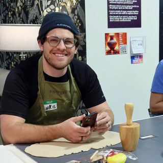 A smiling potter in a navy beanie holds a phone beside a rolled out clay slab and a wooden tool at The Clay Hole in Draper, Utah