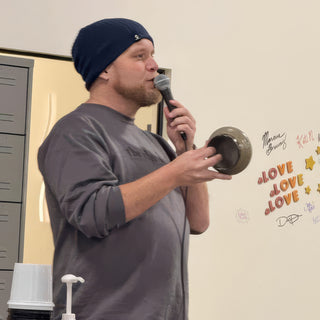 A member in a navy beanie speaks into a microphone while holding up a round bowl with a dark interior at The Clay Hole in Draper, Utah