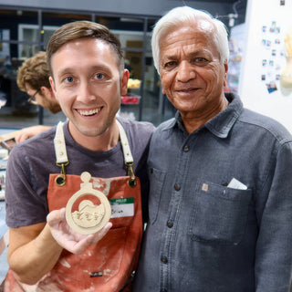 Two friends smiling together while one holds up a small round clay ornament with a cut-out mountain scene at The Clay Hole in Draper, Utah