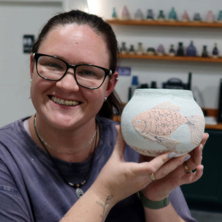 A smiling potter holds up a round pale green pot with a peach fish and dark scale lines at The Clay Hole in Draper, Utah