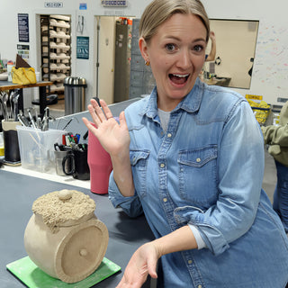 A potter in a denim shirt waving with an excited look beside a round unglazed jar with a rough lid at The Clay Hole in Draper, Utah