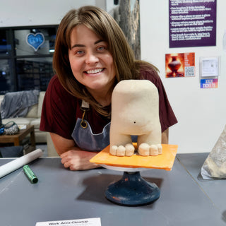 A smiling potter leans beside a rounded cream clay figure with chunky toes, set on an orange board at The Clay Hole in Draper, Utah