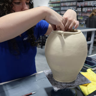 A potter reaching over a large rounded clay pot to pinch and shape its rim on a dark board at The Clay Hole in Draper, Utah